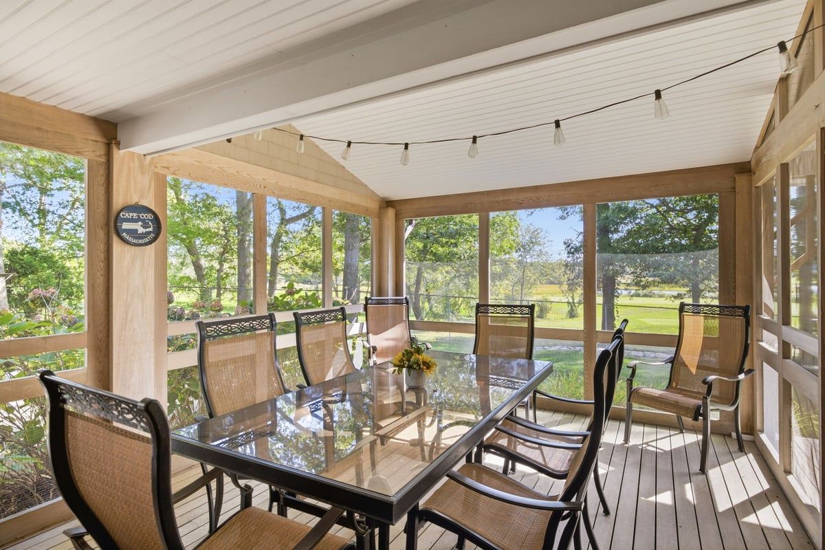A covered outdoor dining area with a glass table and wicker chairs, surrounded by wooden beams and large windows.