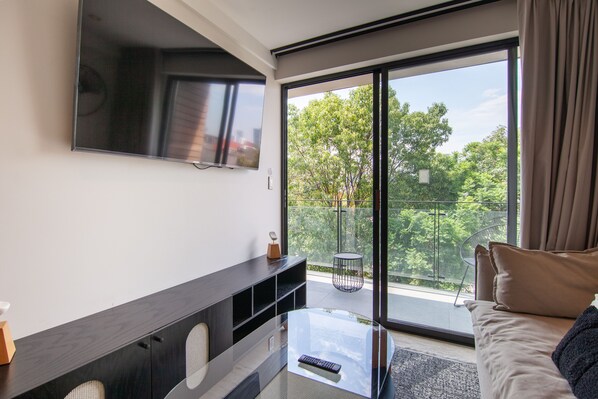 A modern living room with a flat-screen TV mounted on the wall, a glass coffee table, a sofa, and a balcony with a view of trees and a railing. - Zen Charm in Heart of Roma: Unique Osaka-Style Apt (Ciudad de México)
