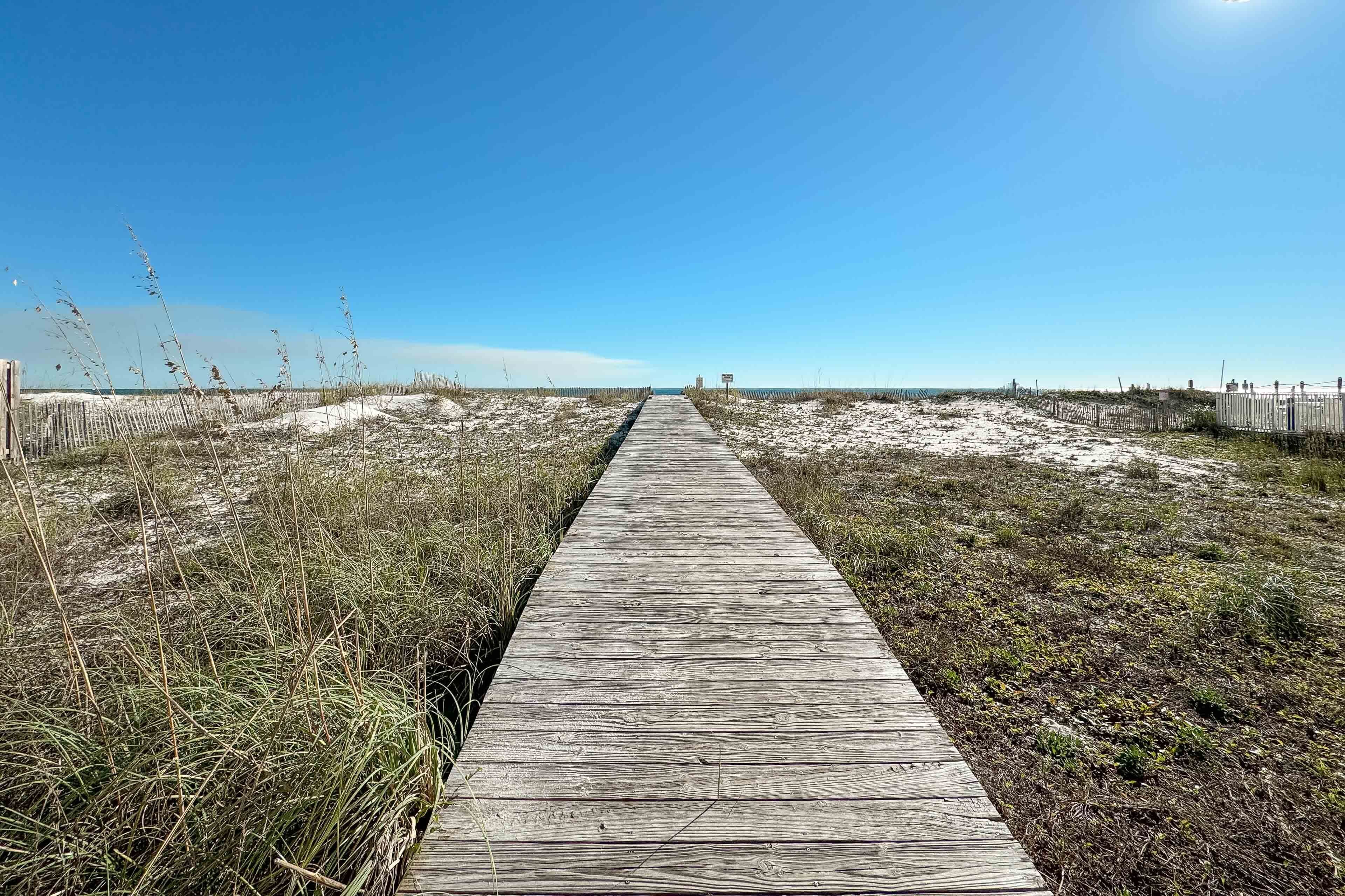 Ein hölzerner Steg führt zu einem Strand mit spärlicher Vegetation und blauem Himmel.