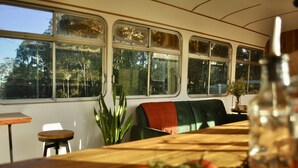 A dining area with a wooden table, a bench, and a view through large windows. - Coffee Grounds - The Bus (Coffee Camp)