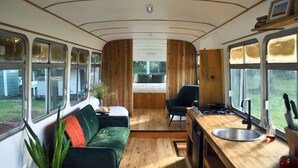 A compact interior with a sofa, a small table, a chair, a kitchenette, and large windows offering views of the outdoors. - Coffee Grounds - The Bus (Coffee Camp)