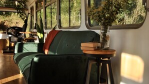 A green couch with a vase of flowers on a stool, a steering wheel, and a window with a view of trees. - Coffee Grounds - The Bus (Coffee Camp)