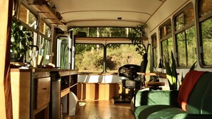 A compact interior with a kitchenette, a dining area, and a seating space with a view of greenery outside. - Coffee Grounds - The Bus (Coffee Camp)