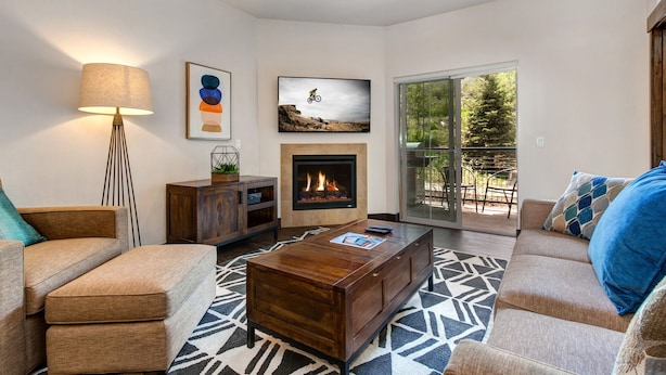 A living room with a fireplace, sofa, coffee table, and a view through a sliding glass door to an outdoor patio. - Gym  Hot Tub & Seasonal Outdoor Pool  Bike Trail  Parking Garage (Park City)