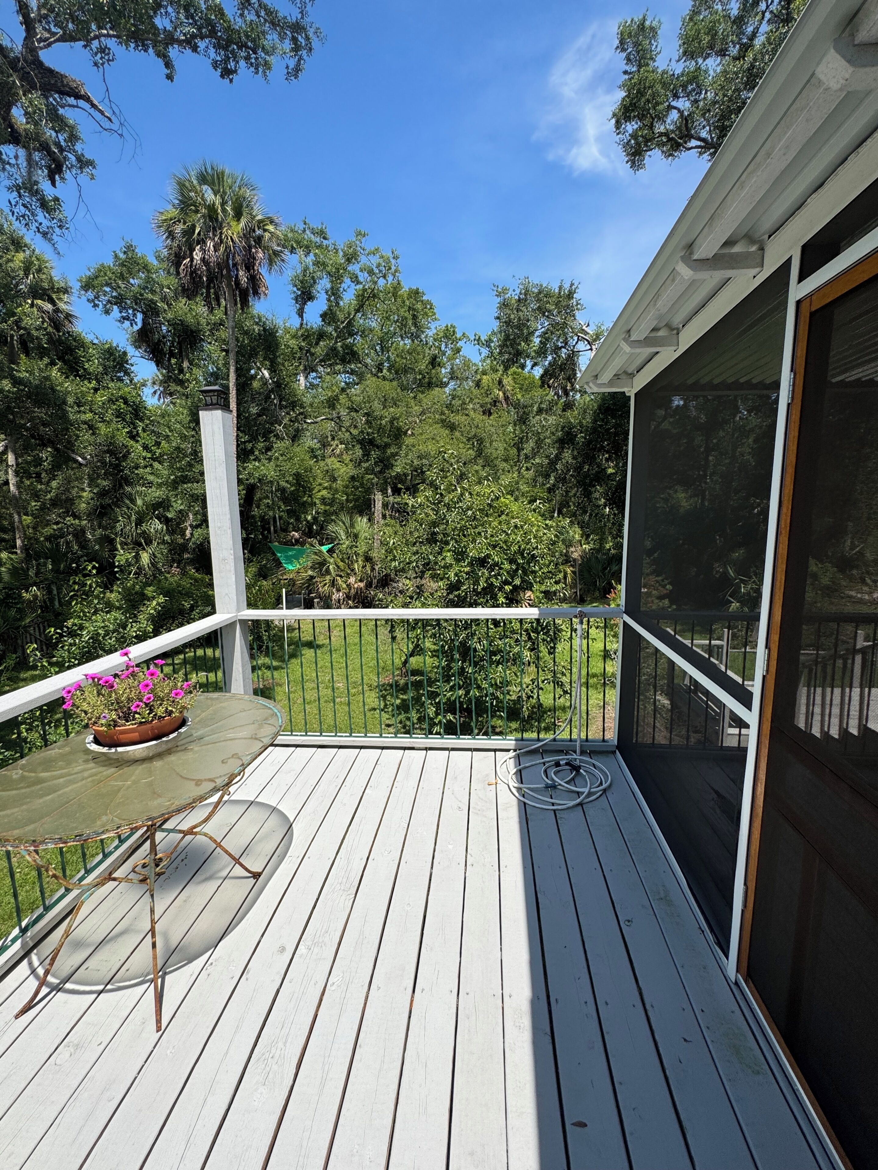 A wooden deck with a railing, a potted plant with pink flowers, and a view of a lush green landscape.