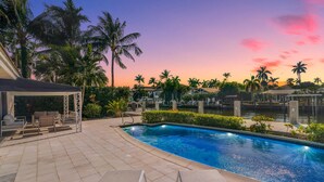 A pool area with lounge chairs and a covered seating area, surrounded by palm trees and a view of a body of water.