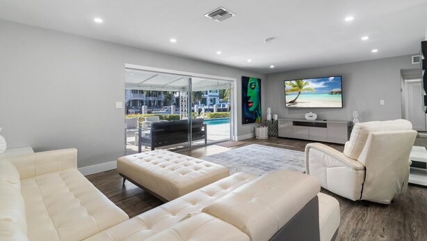 A modern living room with a large glass door leading to a balcony, a flat-screen TV mounted on the wall, and a pool visible through the glass.