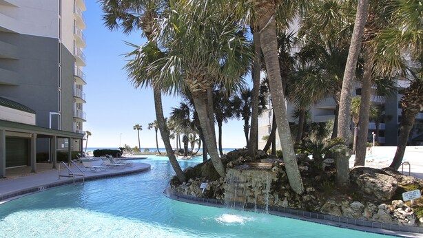Un espace piscine avec une eau d’un bleu limpide, des palmiers et un bâtiment doté de balcons.