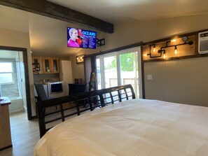 A bedroom with a large bed, a television displaying a music event, a kitchen area, and a sliding glass door leading to an outdoor deck.