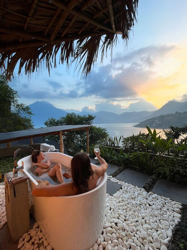Ein Whirlpool mit zwei entspannten Personen, umgeben von einem Kiesgarten mit Blick auf einen See und Berge im Hintergrund.
