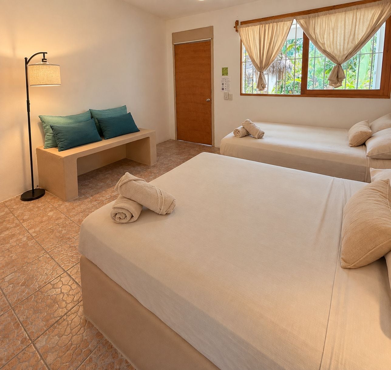 Habitación de hotel con dos camas, un banco, una lámpara de pie y una ventana con cortinas.