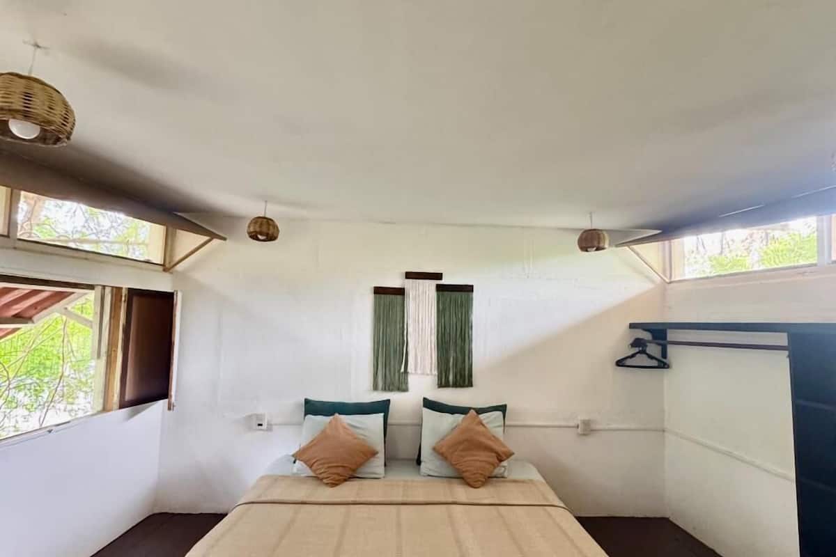 Een eenvoudige, nette kamer met een bed, twee kussens, een raam met gordijnen en een hanglamp.