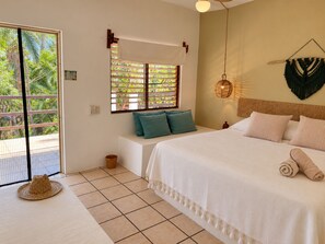 A bedroom with a bed, pillows, a window with a view of greenery, a woven wall hanging, and a woven hat on the floor. - El Eden Escondido (Puerto Escondido)