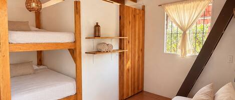 Ein kleines Zimmer mit Etagenbett, einem Einzelbett, einem Fenster mit Vorhängen und einer Holzbalkendecke.