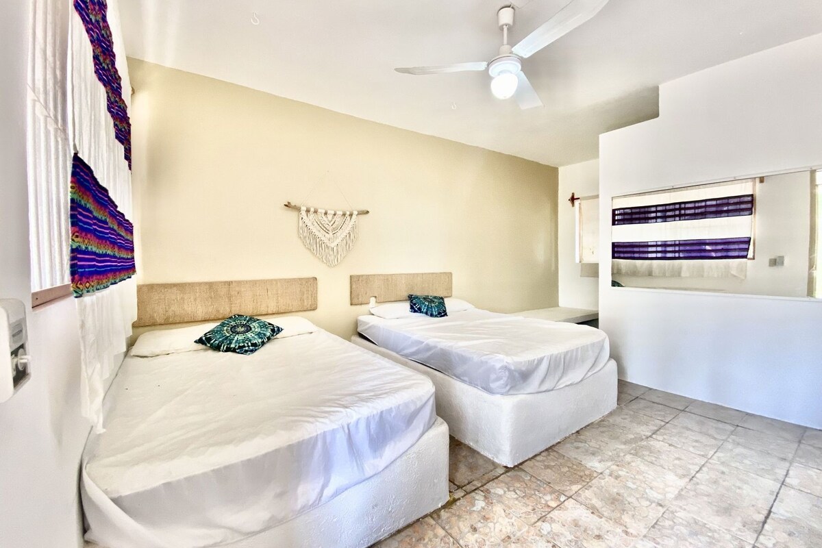 Une chambre d’hôtel avec deux lits, un ventilateur de plafond, une décoration murale en macramé et un grand miroir.