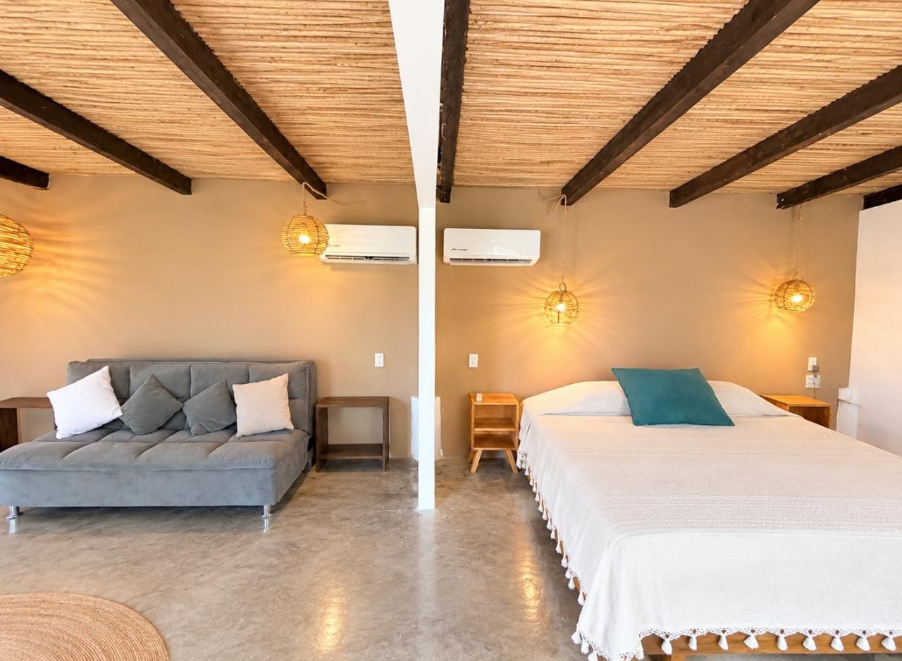A room with a grey sofa, a bed with a white and teal pillow, wooden beams on the ceiling, and two wall-mounted air conditioners.