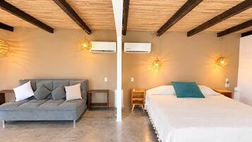 A room with a grey sofa, a bed with a white and teal pillow, wooden beams on the ceiling, and two wall-mounted air conditioners.