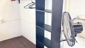 Ein Eckzimmer mit einem dunklen Holzregal, einer Kleiderstange und einem Wandventilator.