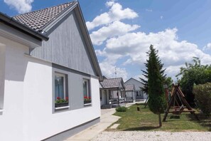 Une rangée de maisons modernes à un étage, avec des toits gris et des murs blancs, alignées les unes à côté des autres, avec une allée en gravier et une balançoire en bois en premier plan.