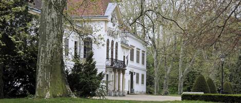 A white building with columns and a balcony, surrounded by trees and a grassy area.