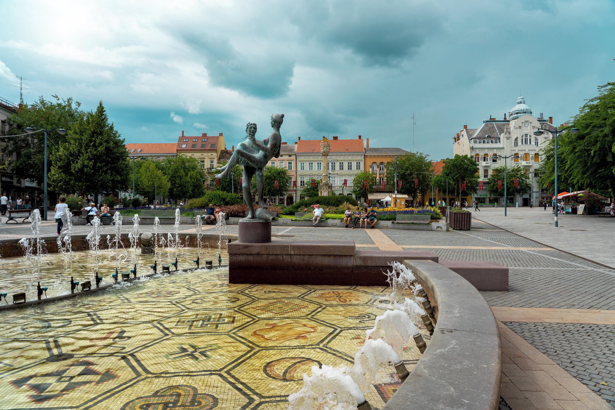Une place publique avec une fontaine, une statue et, en arrière-plan, des bâtiments historiques.