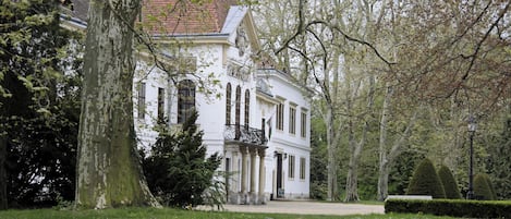 A white building with columns and a balcony, surrounded by trees and a grassy area.