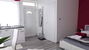 Ein modernes Hotelzimmer mit einem Bett, einem Schreibtisch und einem Sessel. Eine Tür führt in einen weiteren Raum, und ein Fenster mit Vorhängen ist vorhanden.