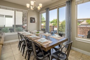 Un espace repas avec une longue table dressée pour six personnes, entourée de chaises, et offrant une vue sur un jardin et un bâtiment à l’extérieur.