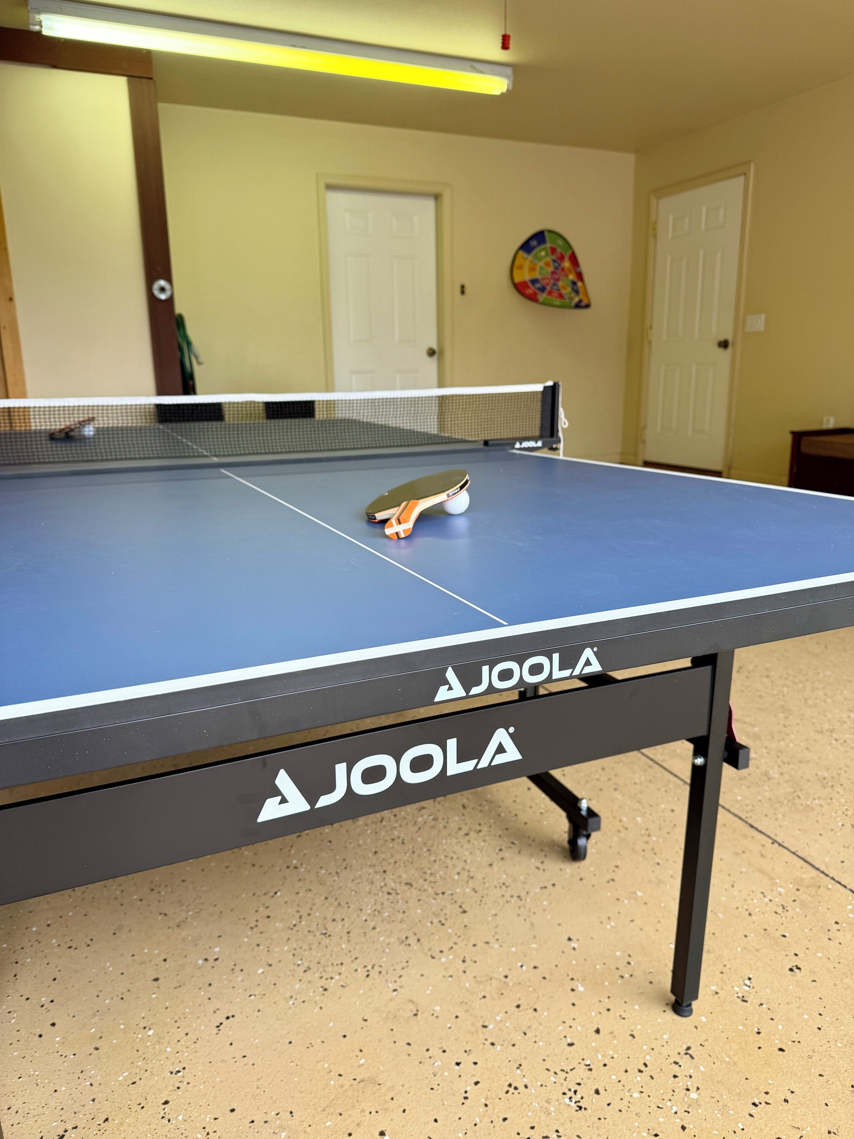 Une table de ping-pong de la marque « Joola », installée à l’intérieur, avec des portes et une décoration murale colorée en arrière-plan.