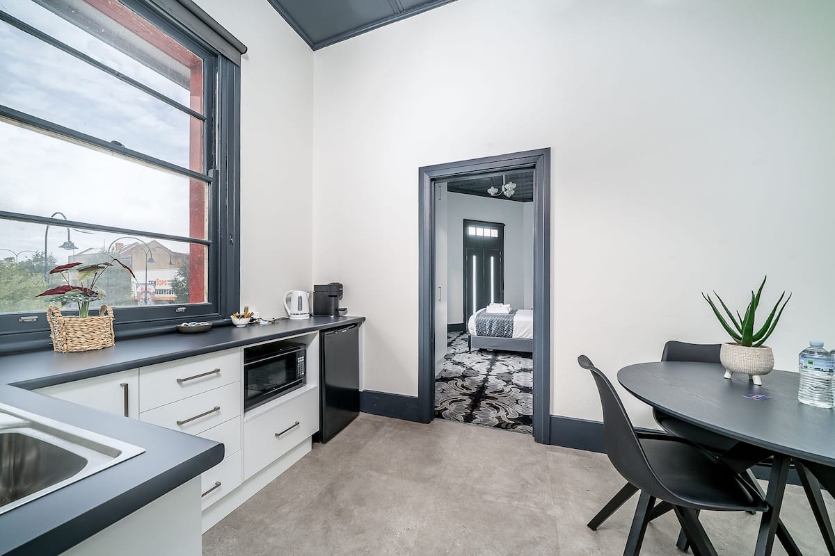 A modern kitchen with a large window, a dining area with a table and chairs, and a view of a bedroom through a doorway.