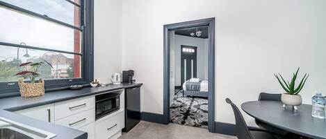 Eine moderne Küche mit einem großen Fenster, eine Essecke mit Tisch und Stühlen sowie ein Blick in ein Schlafzimmer durch einen Durchgang.