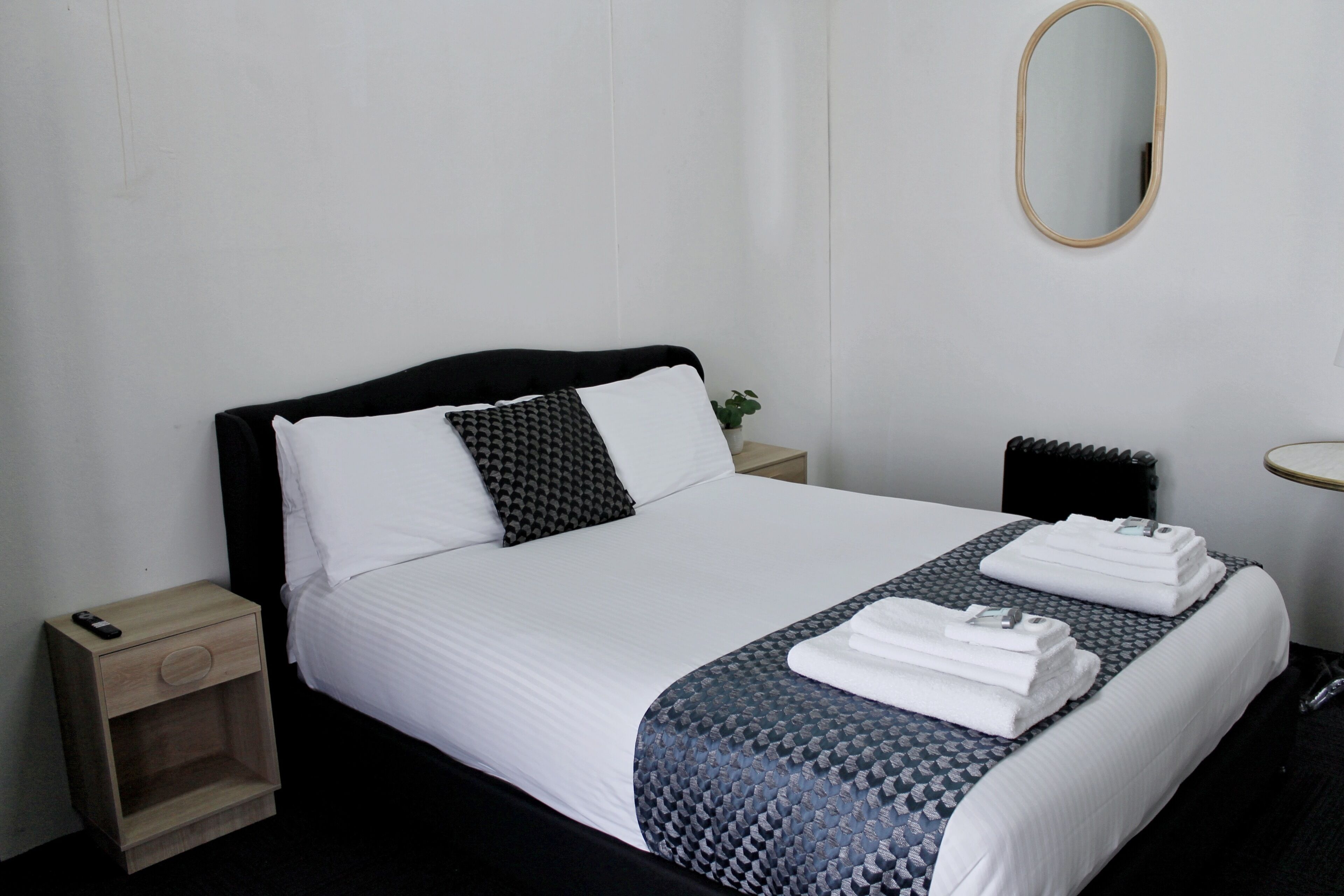 A neatly made bed with white linens, a dark headboard, and a patterned bedspread. There is a small wooden nightstand with a drawer and a remote control on it. A small potted plant is visible on the nightstand.