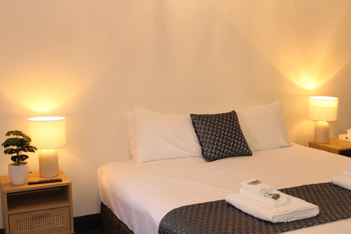 A hotel room with a neatly made bed, two bedside lamps, a small table with a plant, and a bedside table with a remote control.
