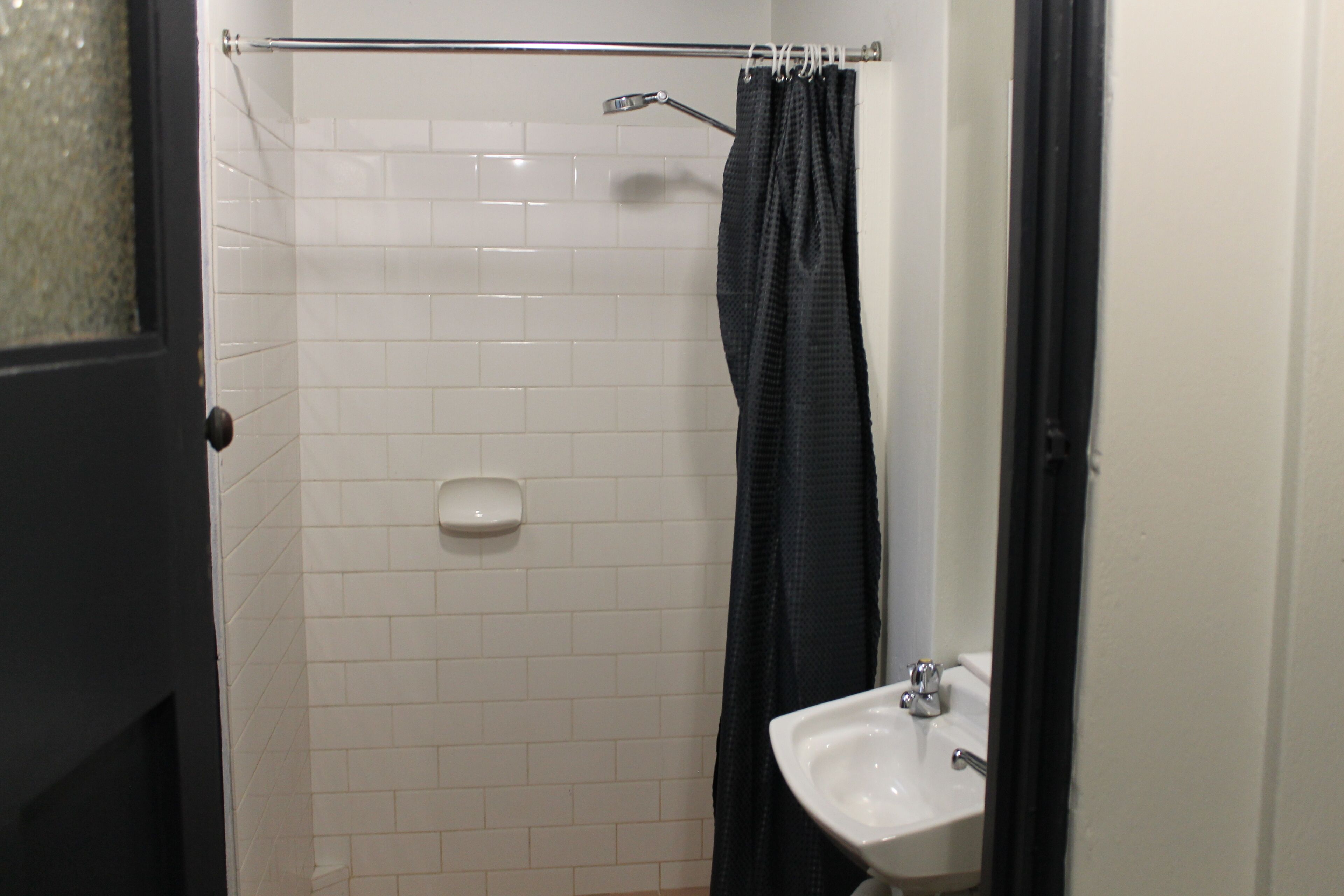 Een badkamer met een witte betegelde muur, een zwart douchegordijn, een wit wastafelmeubel en een glazen deur.