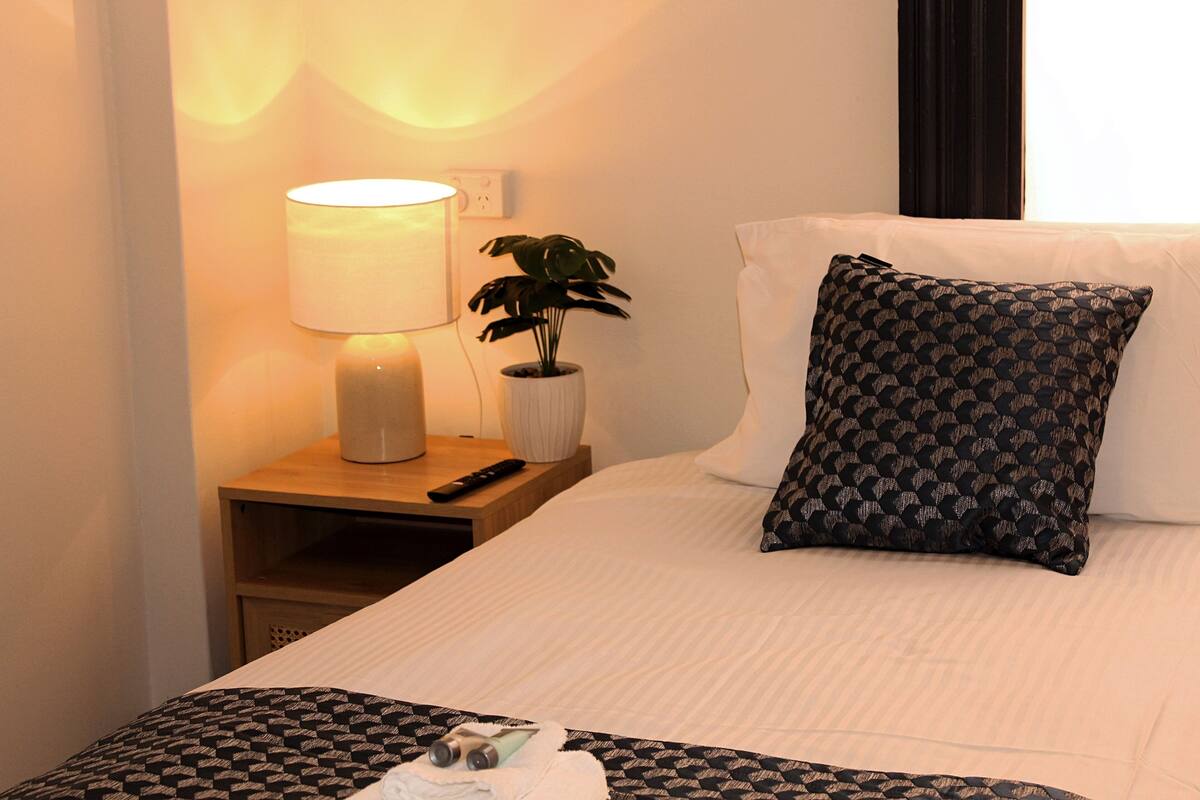 A hotel room with a bed, a nightstand with a lamp, a remote control, a potted plant, and a patterned pillow.
