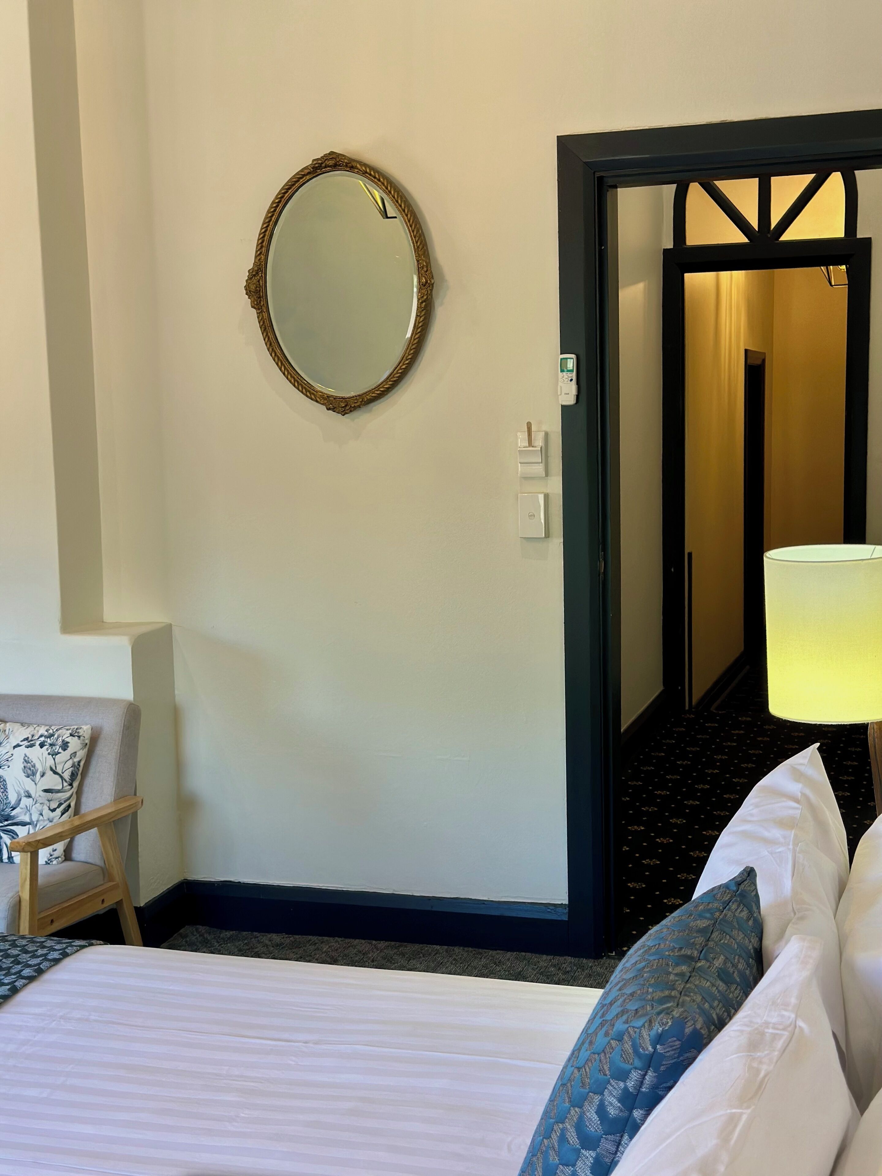 A hotel room with a bed, a bedside table with a lamp, a chair, a wall-mounted mirror, and a door leading to another room.