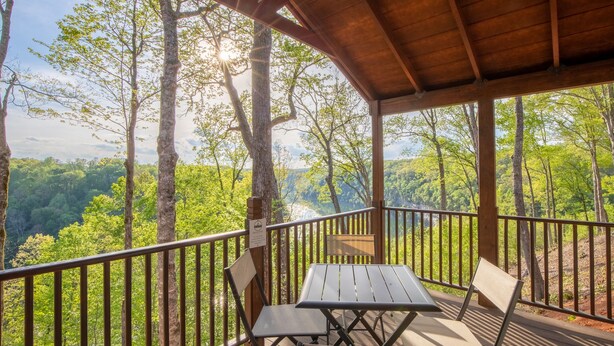 Un espace extérieur abrité, doté d’une table et de chaises, entouré d’une terrasse en bois et d’une rambarde en métal, et offrant une vue sur un paysage boisé.