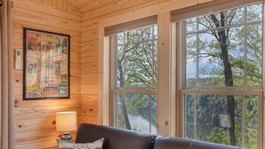 L’intérieur d’une cabane avec un mur en bois, un guide de camping encadré, un canapé et une fenêtre donnant sur une zone boisée.