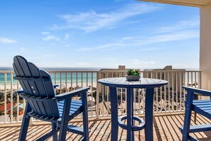 Un balcón con dos sillas Adirondack y una mesita pequeña, con vista a una playa y al océano.
