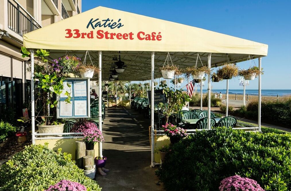 A café with outdoor seating, covered by a canopy, and surrounded by greenery.