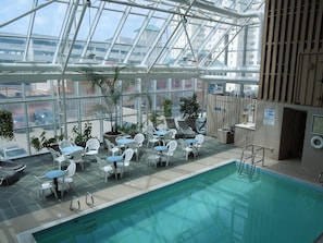 An indoor swimming pool with lounge chairs and tables, surrounded by glass walls and potted plants. - Four Sails - Oceanfront & City View Balconies! (Virginia Beach)