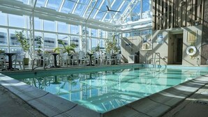 An indoor swimming pool with clear glass walls, surrounded by white chairs and potted plants. - Four Sails - Oceanfront & City View Balconies! (Virginia Beach)