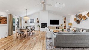 Una sala de estar moderna con un sofá gris, una mesa de comedor y sillas. Hay un televisor montado en la pared y un ventilador de techo.