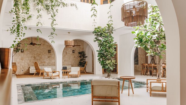 Amplia zona de piscina interior con varios rincones de descanso, incluyendo sillones y una mesa, rodeada de arcos y plantas.
