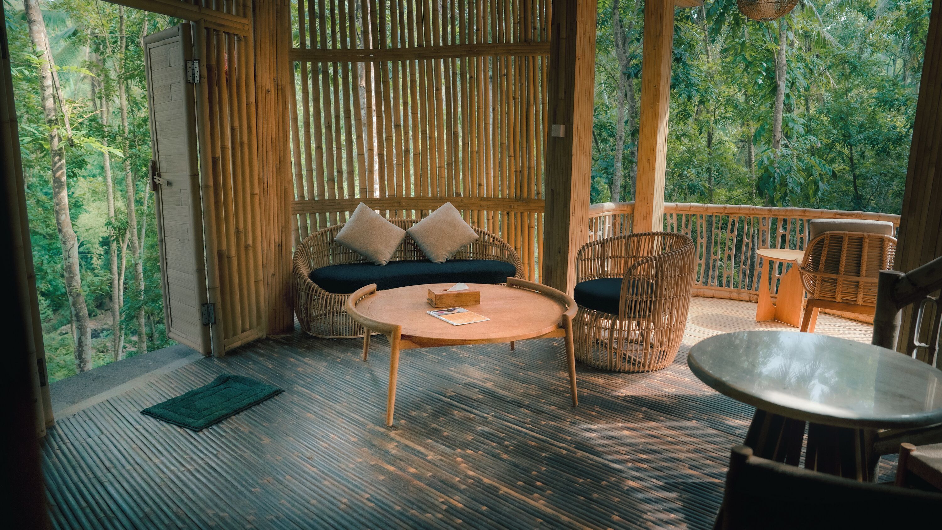 Terraza de madera con un sofá, sillas y una mesa, rodeada de paredes de bambú y vista a un bosque.