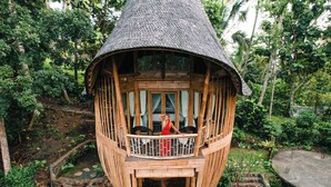 Uma estrutura de bambu com telhado de palha, uma varanda e uma escadaria de pedra que leva a um nível inferior.