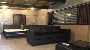 A modern living room with a black sofa, a bed with a mattress, a glass coffee table, and a window with blinds. - Villa Gazamani (Yaese)