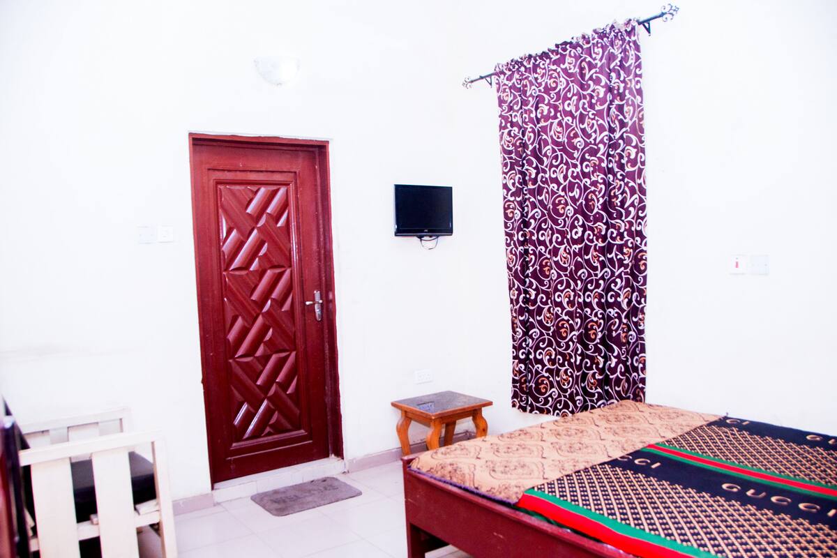 Un dormitorio con una puerta de madera, una mesita de noche pequeña, una cama con ropa de cama estampada y un televisor instalado en la pared.