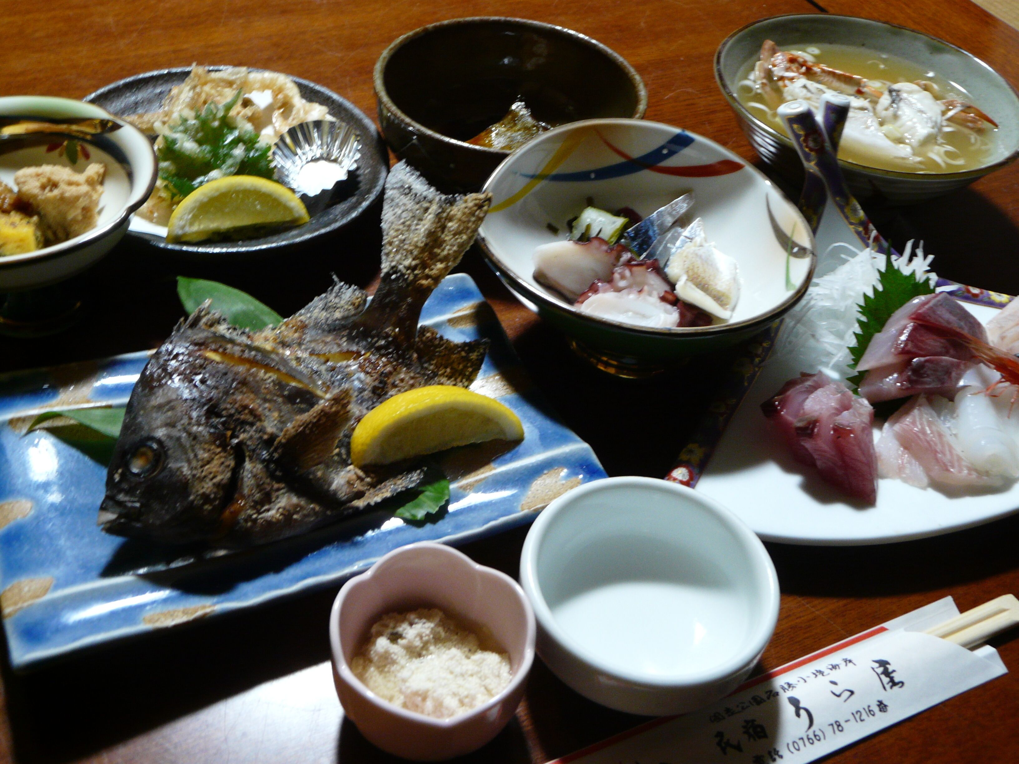 A variety of dishes including a whole fish, sashimi, and a bowl of soup, suggesting a Japanese dining experience.