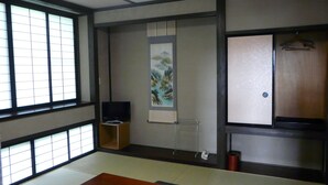 Ein traditioneller japanischer Raum mit Tatami-Bodenbelag, einem niedrigen Holztisch, einem Fernseher, einem Kleiderschrank und einem Bild an der Wand.